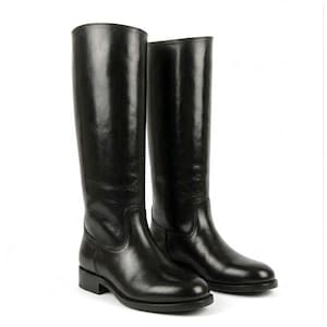 Puede incluir: Un par de botas altas de cuero negro con un acabado liso y pulido, punta redondeada y tacón bajo de bloque. Las botas presentan una silueta limpia y estructurada con costuras visibles a lo largo del empeine y la caña. Logo Morar Leather Gear visible.