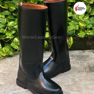 May include: A pair of tall, black leather riding boots. The boots have a smooth, glossy finish and a classic design. The interior of the boots is a light brown color. The boots are displayed on a stone surface.