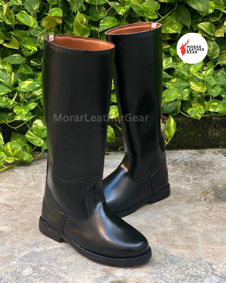 May include: A pair of tall, black leather riding boots. The boots have a smooth, glossy finish and a classic design. The interior of the boots is a light brown color. The boots are displayed on a stone surface.