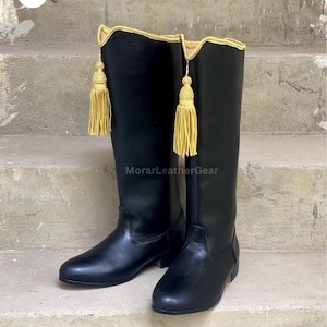 Puede incluir: Un par de botas negras hasta la rodilla con un borde dorado en la parte superior y una borla decorativa dorada colgando del lateral de cada bota. Las botas tienen un acabado liso y tacón bajo, con el texto MorarLeatherGear.