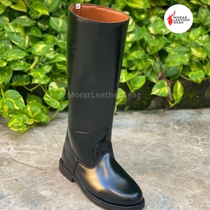 May include: A tall, black leather riding boot with a smooth, glossy finish. The boot has a rounded toe and a brown interior. The boot is approximately 18 inches tall. The background features green foliage.