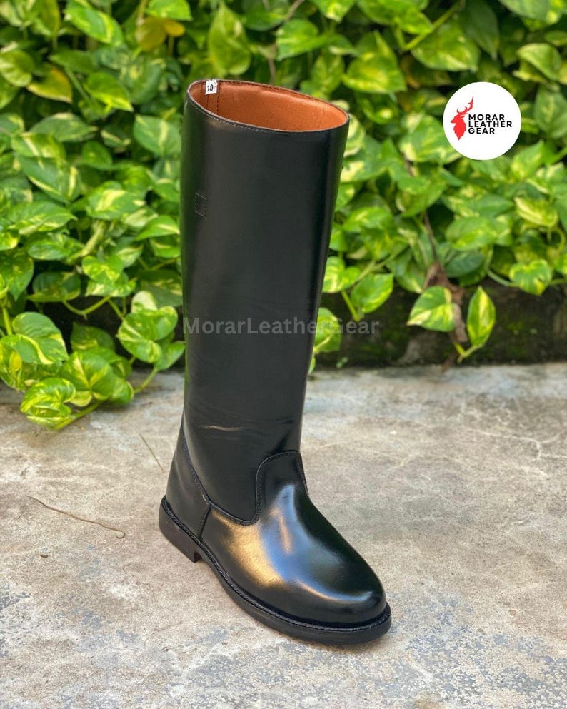 May include: A tall, black leather riding boot with a smooth, glossy finish. The boot has a rounded toe and a brown interior. The boot is approximately 18 inches tall. The background features green foliage.