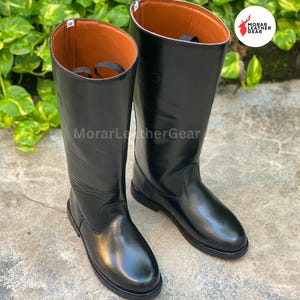 May include: A pair of black leather riding boots with a glossy finish. The boots have a tall shaft, a rounded toe, and a brown interior. The brand name "MorarLeatherGear" is visible in the image.