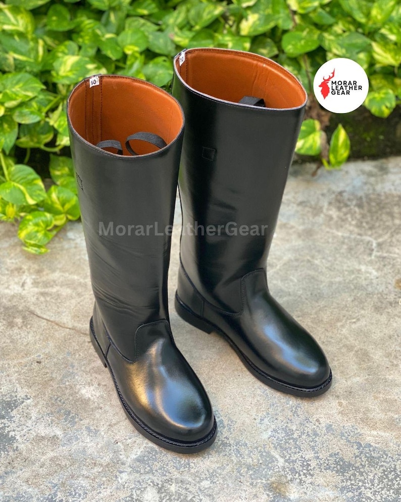May include: A pair of black leather riding boots with a glossy finish. The boots have a tall shaft, a rounded toe, and a brown interior. The brand name "MorarLeatherGear" is visible in the image.