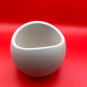 May include: A small, light gray, spherical bowl with a rounded opening. The bowl is made of a smooth, matte material and sits against a bright red background. The bowl's design is simple and modern, suitable for various uses.