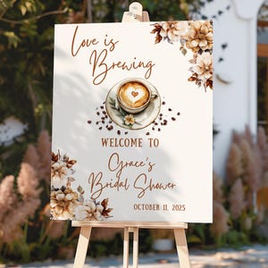 May include: A white sign on a wooden easel with the text "Love is Brewing" and "Welcome to Grace's Bridal Shower October 11, 2025." The sign features a coffee cup with a heart design and floral accents.