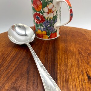 Op de afbeelding: Een zilveren lepel met een bloemenontwerp en een witte mok met een kleurrijk bloemenpatroon. De mok heeft rode, oranje, gele en paarse bloemen. De lepel ligt op een houten oppervlak.