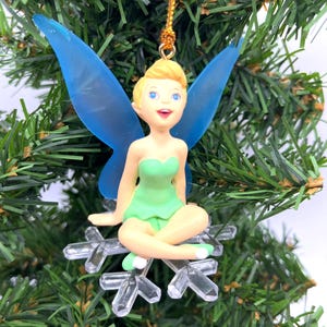 May include: A Christmas tree decoration depicting a fairy with blonde hair, blue wings, and a green dress. The fairy sits on a clear snowflake. The ornament is suspended by a gold string, set against a backdrop of green Christmas tree branches.