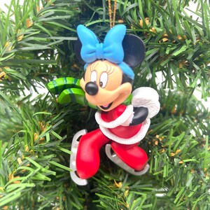 May include: A Minnie Mouse Christmas ornament. Minnie is wearing a red outfit with white trim, a blue bow, and ice skates. The ornament is hanging on a green Christmas tree.