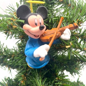 May include: A Mickey Mouse Christmas tree ornament. The ornament features Mickey Mouse in a blue outfit, playing a brown violin. The ornament has black ears, a pink face, and white gloves. A gold string is attached for hanging.