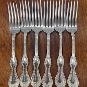 May include: Six silver forks with ornate handles are arranged on a dark wooden surface. The forks have tines and decorative detailing on the handles. The silverware is likely for a formal dining setting.