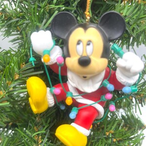 May include: A Christmas tree ornament featuring Mickey Mouse in a Santa suit, tangled in colorful Christmas lights. The ornament has black ears, a yellow face, and yellow shoes. The background is a green Christmas tree.