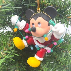 May include: A Christmas tree ornament featuring Mickey Mouse in a Santa Claus outfit, holding a string of colorful lights. The ornament has black ears, a red suit with white trim, yellow shoes, and a gold hanging string.
