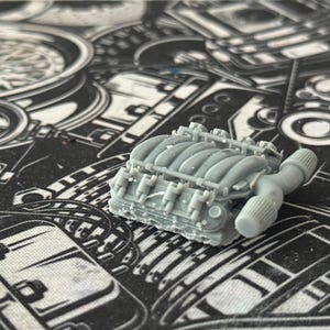 Peut inclure: Un modèle réduit gris clair d'un moteur, avec des détails précis des cylindres, des soupapes et du collecteur d'échappement. Le modèle est posé sur un fond à motifs noir et blanc.