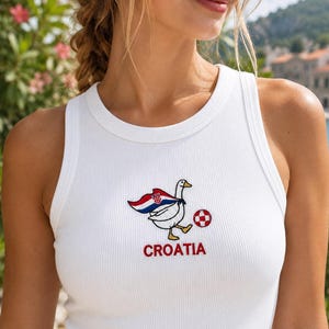 May include: A white ribbed tank top featuring an embroidered graphic of a goose wearing a cape with the Croatian flag design, kicking a red and white checkered soccer ball, with the word CROATIA embroidered in red lettering underneath the goose.