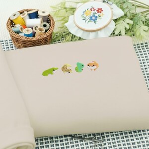 May include: A beige notebook with four colorful reptile-shaped stickers. A green frog, a yellow and brown gecko, a green lizard, and an orange and brown gecko. A basket of thread spools and an embroidered hoop are in the background.