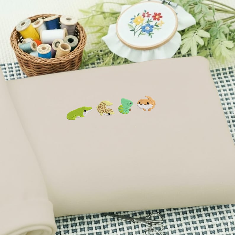 May include: A beige notebook with four colorful reptile-shaped stickers. A green frog, a yellow and brown gecko, a green lizard, and an orange and brown gecko. A basket of thread spools and an embroidered hoop are in the background.