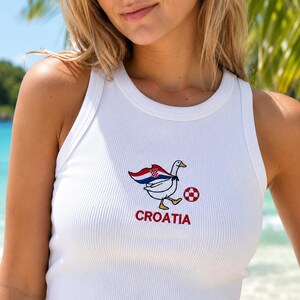May include: A white ribbed tank top featuring an embroidered graphic of a white goose wearing a cape with the red, white, and blue Croatian flag pattern. The goose is kicking a red and white checkered soccer ball, with the word CROATIA in red.