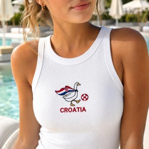 May include: A white ribbed tank top featuring an embroidered graphic of a white goose wearing a cape with the Croatian flag design, kicking a red and white checkered soccer ball, with the word CROATIA embroidered in red lettering below the goose.