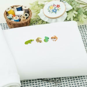 May include: White fabric with four colorful reptile-shaped embellishments. A green frog, a tan gecko, a green lizard, and a brown lizard are displayed. A basket of thread spools and an embroidery hoop with floral design are in the background.