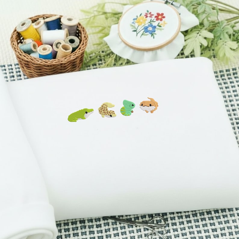 May include: White fabric with four colorful reptile-shaped embellishments. A green frog, a tan gecko, a green lizard, and a brown lizard are displayed. A basket of thread spools and an embroidery hoop with floral design are in the background.