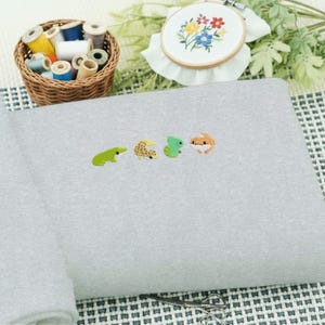 May include: A light gray sweatshirt embroidered with four small, colorful reptile designs: a green frog, a tan gecko, a green lizard, and an orange gecko. A basket of thread spools and an embroidery hoop with floral embroidery are in the background.