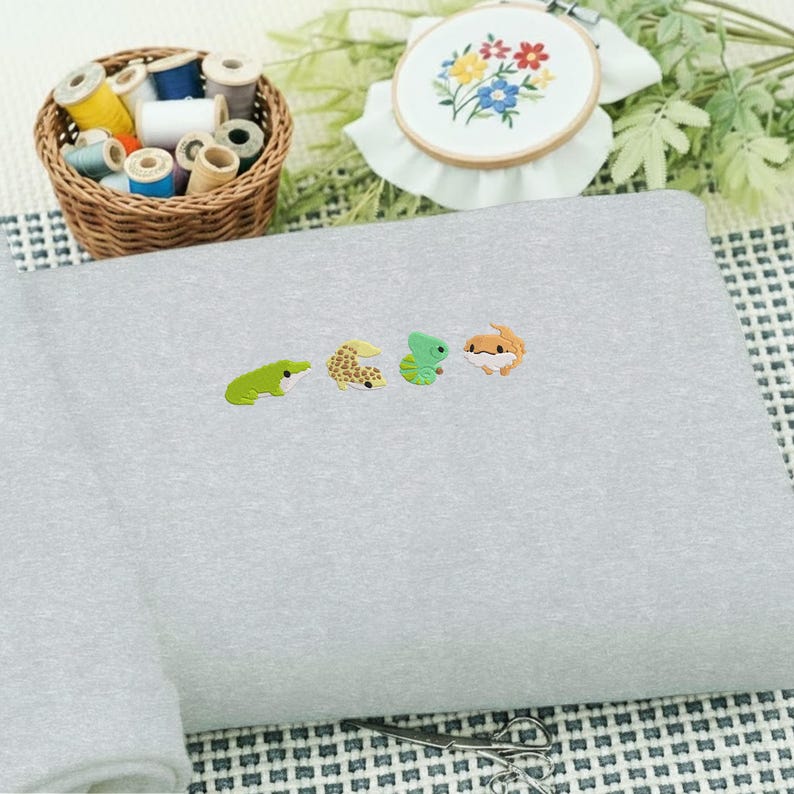 May include: A light gray sweatshirt embroidered with four small, colorful reptile designs: a green frog, a tan gecko, a green lizard, and an orange gecko. A basket of thread spools and an embroidery hoop with floral embroidery are in the background.