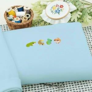 May include: A light blue fabric with four embroidered reptile designs: a green frog, a tan gecko, a green gecko, and an orange lizard. A small basket of thread spools and an embroidery hoop with floral embroidery are in the background.