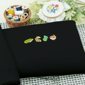 May include: Black fabric with embroidered colorful reptile designs. A frog, gecko, chameleon, and bearded dragon are featured. A basket of thread spools and an embroidered floral hoop are in the background.