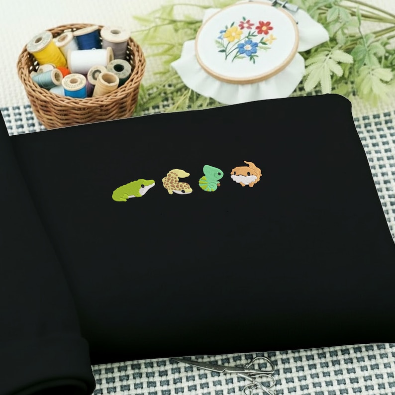 May include: Black fabric with embroidered colorful reptile designs. A frog, gecko, chameleon, and bearded dragon are featured. A basket of thread spools and an embroidered floral hoop are in the background.