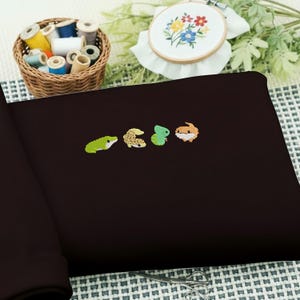 May include: A dark brown fabric with four embroidered reptile designs in green, tan, and orange. A basket of colorful thread spools and an embroidery hoop with a floral design are in the background.