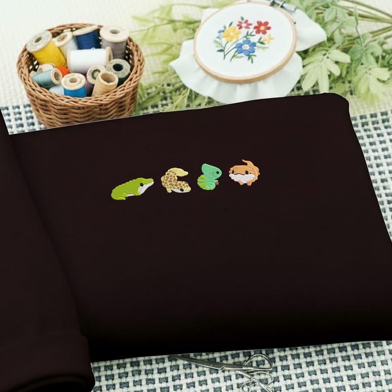 May include: A dark brown fabric with four embroidered reptile designs in green, tan, and orange. A basket of colorful thread spools and an embroidery hoop with a floral design are in the background.