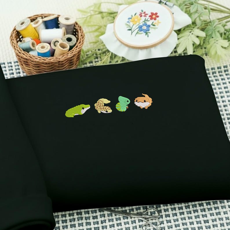 May include: Black fabric with embroidered reptile designs. The embroidered creatures include a green frog, a tan and brown lizard, a green gecko, and a brown and tan lizard. A basket of thread spools and an embroidery hoop with floral embroidery are in the background.