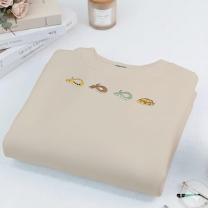 May include: A beige sweatshirt featuring four cartoon snake designs in yellow, brown, green, and yellow-brown. The snakes are arranged horizontally across the chest of the folded garment. The sweatshirt is made of a soft material.