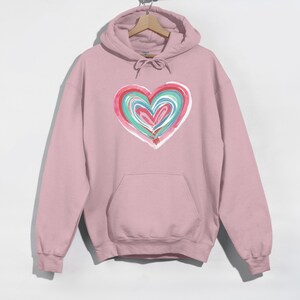 May include: A light pink hoodie with a large heart design on the front. The heart is composed of layered brushstrokes in shades of red, teal, and white. The hoodie has a drawstring hood and a front pocket.