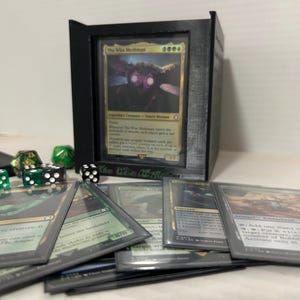 May include: A black card holder with a Magic: The Gathering card featuring "The Wise Mochman" and other cards. Several dice are scattered around the holder. The card holder has the text "The Deck of Many Things" on it.