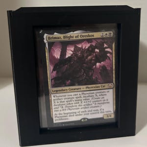 May include: A framed Magic: The Gathering card featuring "Brimaz, Blight of Oreskos." The card has a dark, detailed illustration of a Phyrexian Cat with text describing its abilities. The frame is black, creating a clean presentation.