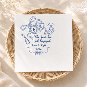 May include: A square white paper napkin featuring a blue ink illustration of two heart-shaped balloons with the letters A and K, a champagne bottle, and a diamond ring. The text reads: The Year We got Engaged, Amy & Kyle, 2026.
