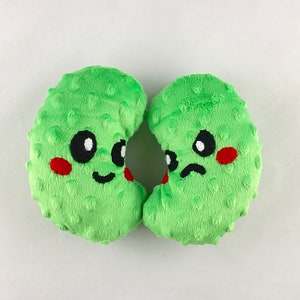 May include: Two green plush toys shaped like half-moons with embroidered faces. One toy has a happy face with black eyes and a red smile. The other toy has a sad face with black eyes and a frown.