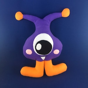 Puede incluir: Un juguete de peluche morado con brazos y piernas naranjas. El juguete tiene un ojo grande blanco con una pupila negra y una pequeña boca rosa con dos dientes blancos.