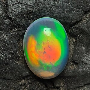 May include: A vibrant, oval-shaped opal gemstone. The opal displays a mesmerizing play of color, with hues of green, orange, and blue swirling within. The gemstone is set against a dark, textured background, highlighting its brilliance.