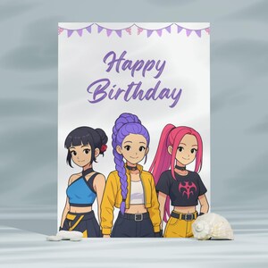 Op de afbeelding: Een verjaardagskaart met drie meisjes in anime-stijl, elk met een unieke haarstijl en outfit. De kaart toont de woorden "Happy Birthday" in paarse letters. Een zeeschelp en kleine witte stenen staan ​​op de voorgrond.