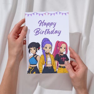 Op de afbeelding: Een verjaardagskaart met een anime-achtige illustratie van drie meisjes. De kaart leest "Happy Birthday" in paarse letters, met een bannerontwerp bovenaan. De meisjes hebben verschillende kapsels en outfits. De kaart wordt door twee handen vastgehouden.