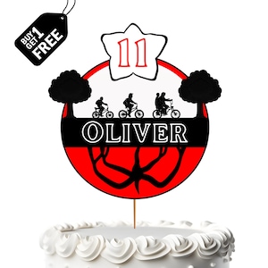 May include: A cake topper with a red and black design featuring silhouettes of people on bikes, the name "OLIVER", and the number "11". The topper is on a white frosted cake. A black tag says "BUY 1 GET 1 FREE".