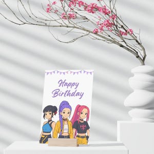 Op de afbeelding: Een verjaardagskaart met de woorden "Happy Birthday" in paarse letters. De kaart toont een illustratie van drie meisjes in anime-stijl. De kaart wordt ondersteund door een klein houten blok. Op de achtergrond staat een witte vaas met roze bloemen.