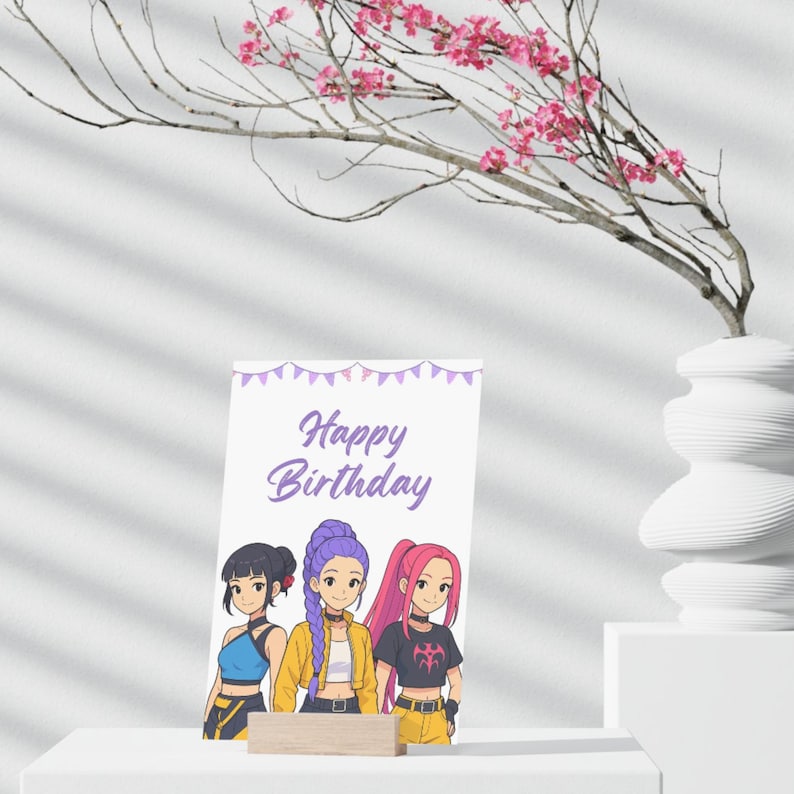 Op de afbeelding: Een verjaardagskaart met de woorden "Happy Birthday" in paarse letters. De kaart toont een illustratie van drie meisjes in anime-stijl. De kaart wordt ondersteund door een klein houten blok. Op de achtergrond staat een witte vaas met roze bloemen.