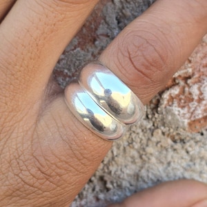 May include: A pair of shiny, silver rings stacked together, reflecting light. The rings are smooth and rounded, creating a simple yet elegant design. The rings are shown on a finger, against a brick background.
