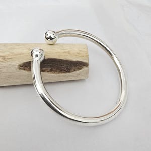May include: A silver-colored, open-ended bangle bracelet. The bracelet has a smooth, curved design with a small, textured ball at each end. The bracelet is resting on a piece of light-colored wood against a white background.
