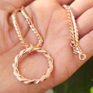 Op de afbeelding: Een roségouden ketting met een cirkelvormige, gevlochten hanger. De ketting is ook roségoud, met een gedraaid patroon. De sluiting is zichtbaar. De ketting wordt op een hand getoond.