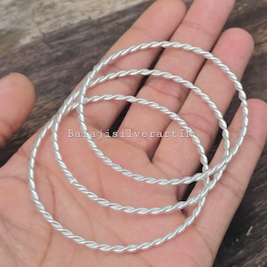 May include: Three silver-colored, twisted-rope design bangles. The bangles are displayed on a hand, with the text "BalajisilverartIN" visible. The bangles are made of a shiny, metallic material.